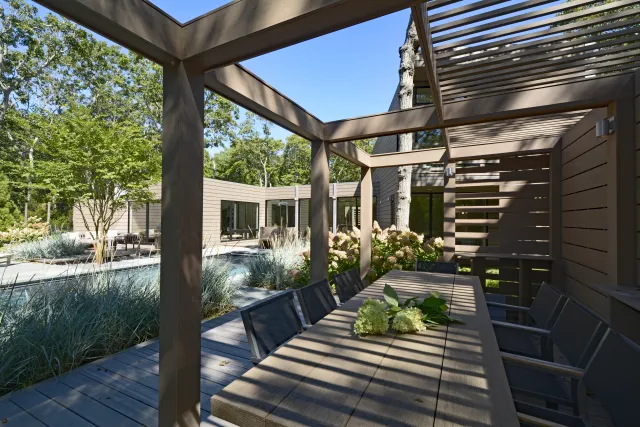 Modern outdoor patio with wooden pergola, dining table, chairs, and surrounding greenery under blue sky.