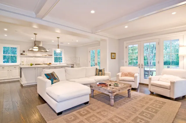 Bright open-concept living room and kitchen with white furniture, wooden floor, and large glass doors to garden.