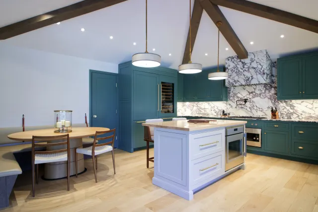 Modern kitchen with teal cabinets, white marble backsplash, central island, round dining table, and wooden ceiling beams.