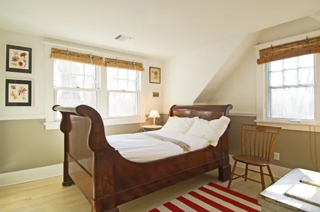 Bright bedroom with antique wooden sleigh bed, white bedding, wooden chair, and striped red rug under natural light