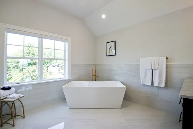 Modern bathroom with a freestanding white bathtub, large window, gold faucet, and towels on the wall.