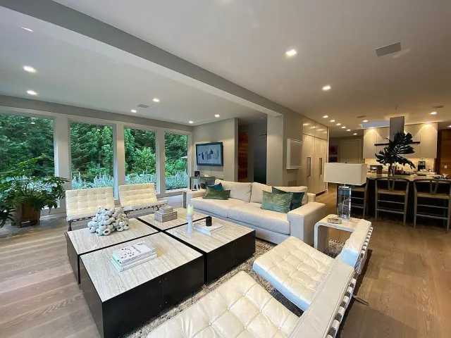Modern living room with cream chairs, beige sofa, marble coffee tables, and large windows overlooking greenery.