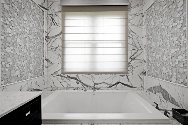 Modern bathroom with marble walls, a large white bathtub, pebble mosaic accents, and a window with a light shade.