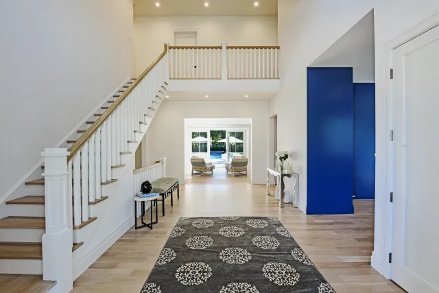 Spacious modern entryway with wooden stairs, patterned rug, white walls, and blue accent wall leading to outdoor seating area.