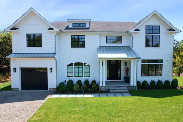 Modern white two-story house with black window frames, a front porch, garage, and manicured lawn under blue sky