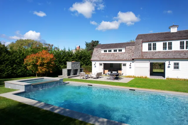 Modern backyard with a clear blue swimming pool, hot tub, outdoor fireplace, and white two-story house under a sunny sky.