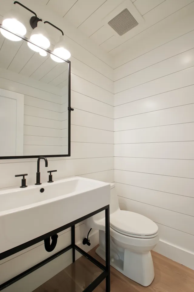 Modern white bathroom with black fixtures, horizontal shiplap walls, and wood-tone flooring under sink and toilet.