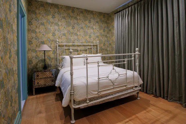 Cozy bedroom with white iron bed, floral wallpaper, wooden floor, bedside table, lamp, and gray curtains.