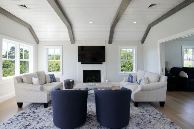 Bright modern living room with vaulted ceiling, large windows, neutral sofas, navy chairs, and a mounted TV above fireplace.