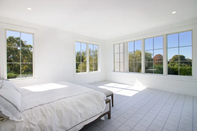 Bright, spacious bedroom with large windows, white walls, and a neatly made bed with white linens.