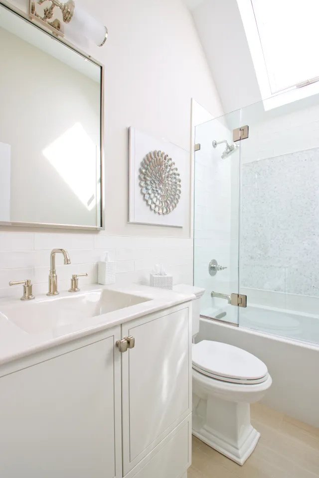 Bright modern bathroom with white vanity, glass shower, and decorative wall art under skylight window.
