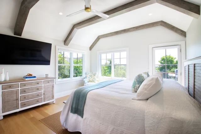 Bright bedroom with vaulted wood beam ceiling, large windows, white bedding, and a wall-mounted TV.