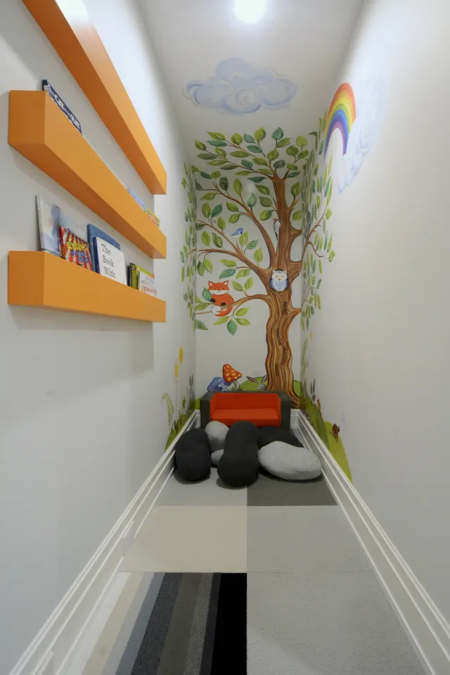 Cozy children's reading nook with wall mural of tree, animals, rainbow, orange shelves with books, stones, and small orange chair.