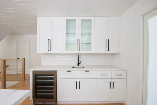 Modern white kitchen with black handles, marble countertop, built-in wine fridge, and glass-front cabinets.
