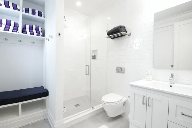 Bright modern bathroom with glass shower, white tiles, floating toilet, and organized shelving with towels.