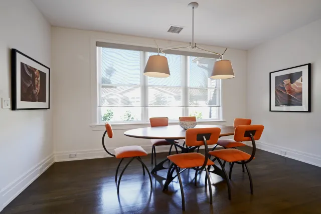 Modern dining room with orange chairs, round wooden table, pendant lights, and framed wall art in bright space