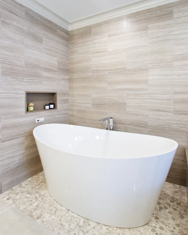 Modern white freestanding bathtub in bathroom with beige tiled walls and pebble stone floor.