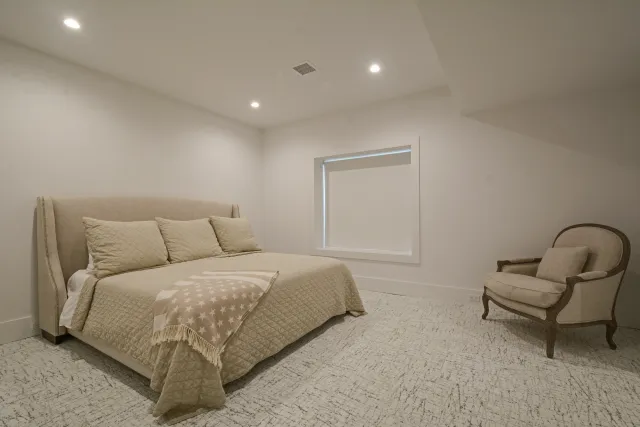 Minimalist bedroom with beige bed, matching armchair, star-patterned throw, and soft lighting under sloped ceiling