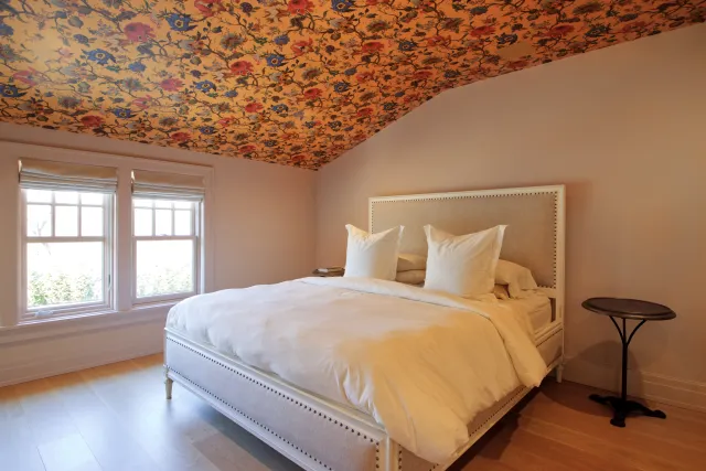Bright bedroom with floral ceiling, large white bed, two windows, and minimalist side table on wooden floor