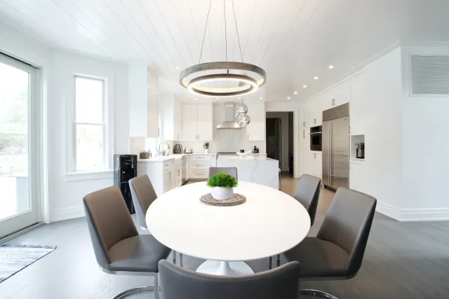 Bright modern kitchen with white cabinets, round dining table, gray chairs, circular chandelier, and stainless steel appliances.