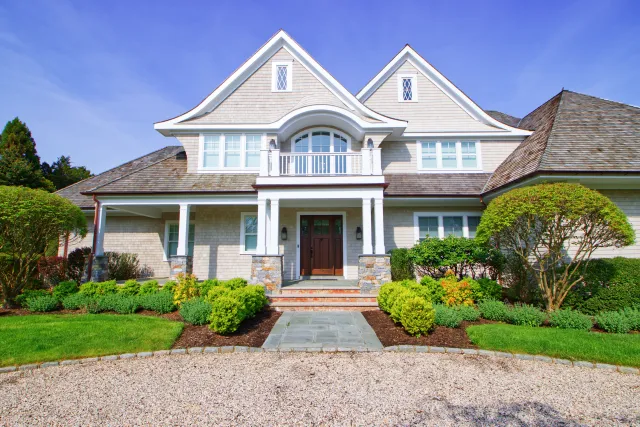Large two-story house with stone porch, symmetrical windows, neatly trimmed bushes, and gravel driveway under blue sky