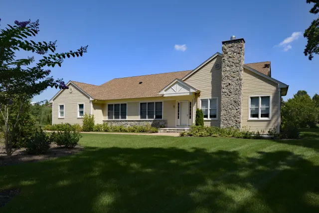 Single-story beige house with a stone chimney surrounded by green lawn and trees under a blue sky.