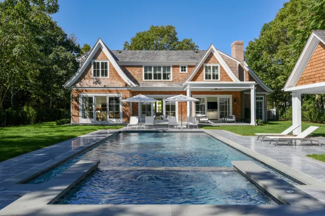 Modern house with large swimming pool, lounge chairs, umbrellas, and lush green trees in the background.