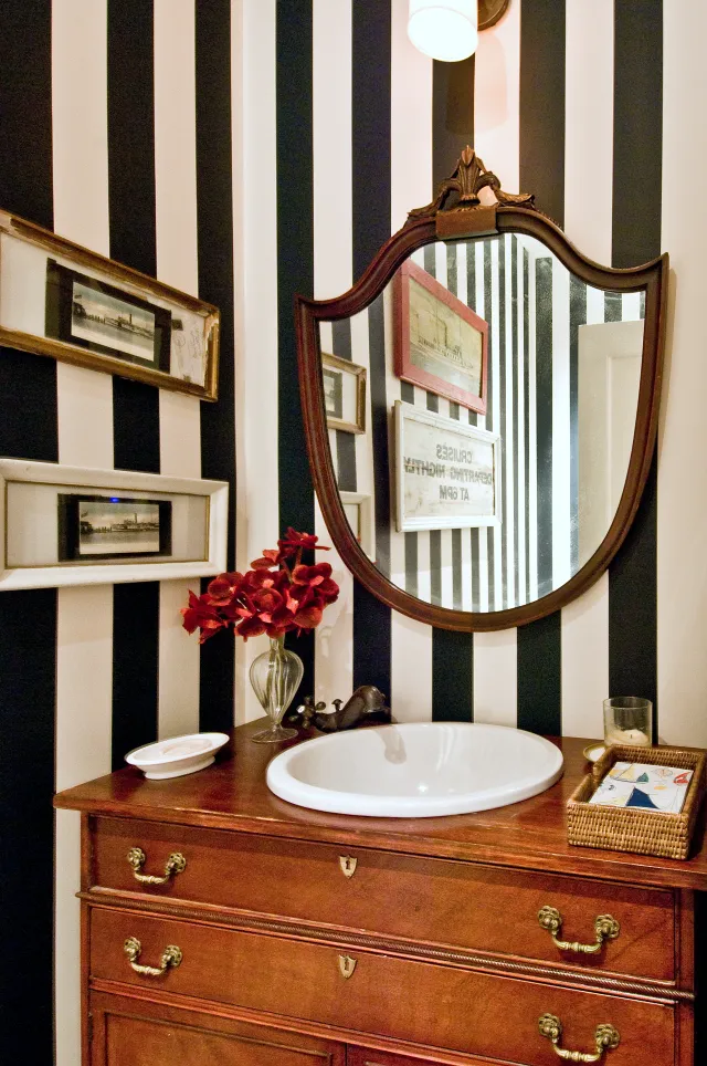 Vintage bathroom vanity with round sink, ornate mirror, striped black and white wall, and red flowers in vase.