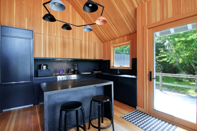 Modern kitchen with black cabinets, wooden walls, island with stools, and large glass door to outdoor greenery.