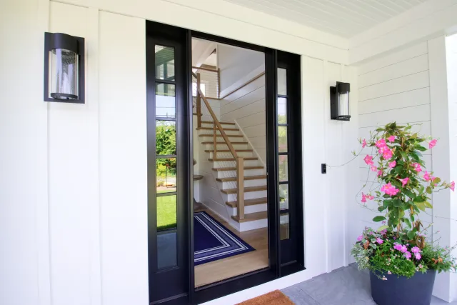 Black framed glass front door with modern wall lights and flowering plant on white porch outside wooden staircase.