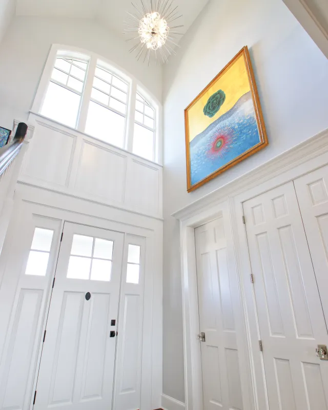 Bright entryway with tall white front door, arched windows, modern chandelier, and colorful framed artwork.