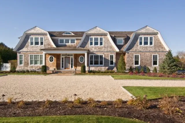 Large shingle-style house with multiple dormers, oval windows, gravel driveway, and well-maintained landscaping under clear sky