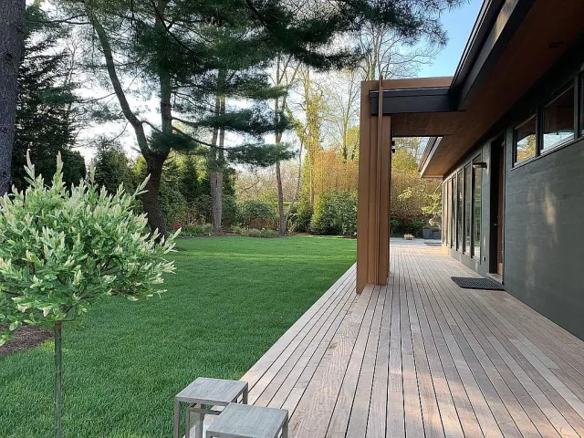 Modern wooden deck extending from house with green lawn, trees, and small potted tree in yard.