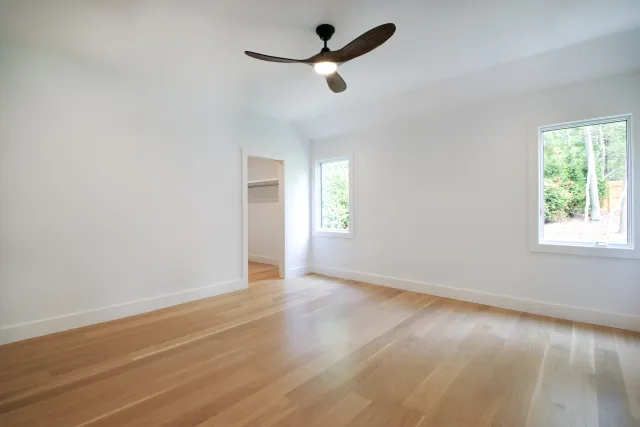 Empty room with light wood floor, white walls, ceiling fan, and two windows showing outdoor greenery.