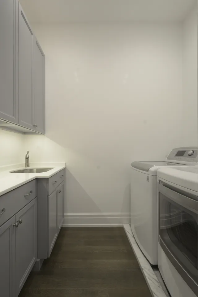 Modern laundry room with gray cabinets, white countertop, stainless steel sink, washer, dryer, and hardwood flooring.