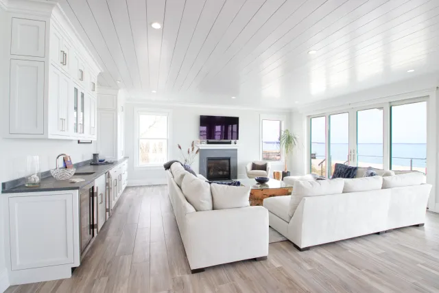 Bright open living room with white sofas, hardwood floors, large windows, fireplace, and coastal view