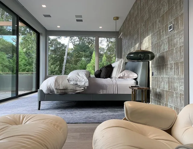 Modern bedroom with large windows, gray bed, textured accent wall, and cream leather chairs in foreground