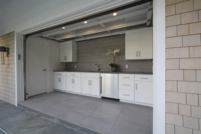 Modern outdoor kitchen with white cabinetry, stainless steel appliances, and gray tile backsplash under a garage door.