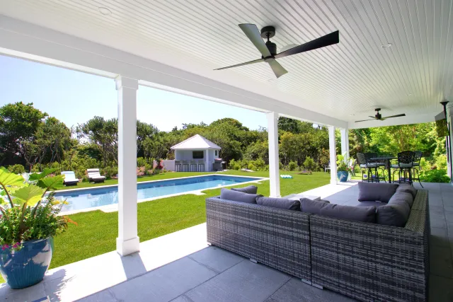 Covered patio with wicker sofa, ceiling fans, and pool view in a green backyard on a sunny day
