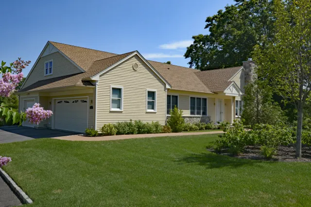 Single-story beige house with attached garage, manicured lawn, blooming trees, and clear blue sky.