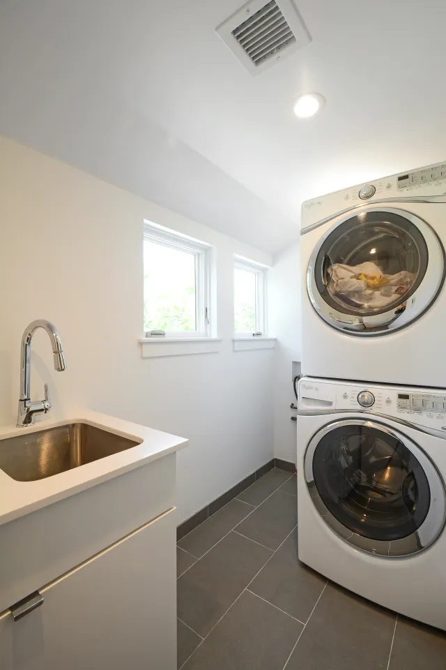 Bright modern laundry room with stacked washer and dryer next to a white sink and two windows.