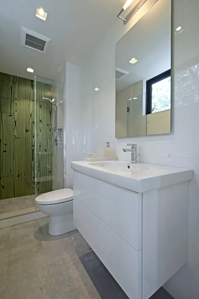 Modern bathroom with a white vanity, large mirror, glass shower with green wood-patterned wall, and gray floor tiles.