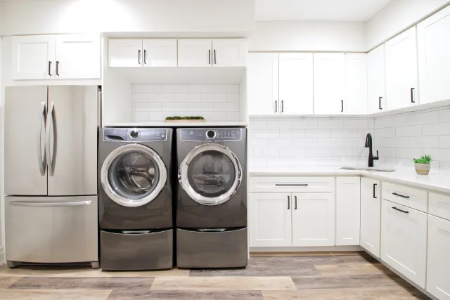 Modern laundry room with front-loading washer and dryer, white cabinets, stainless steel refrigerator, and wood flooring.