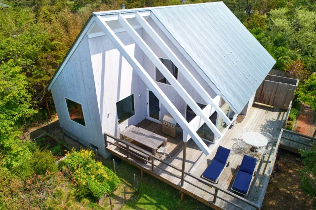 A modern white cabin with large wooden deck, outdoor furniture, and surrounded by green trees in a forest setting.