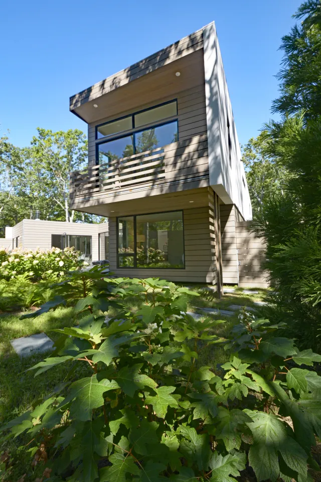 Modern two-story house with large windows and balcony surrounded by lush green plants on a sunny day