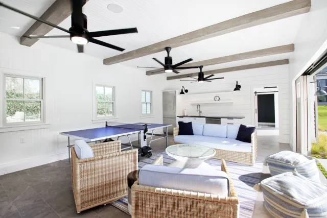 Bright indoor recreational room with wicker furniture, blue cushions, ping pong table, and ceiling fans under wooden beams.