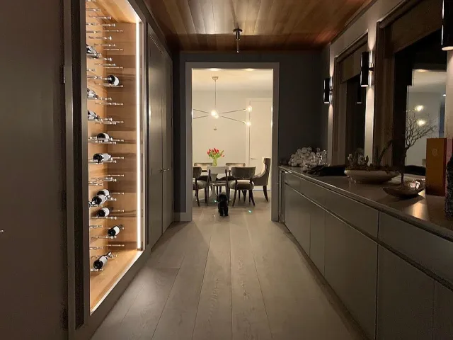 Dimly lit modern hallway with wooden ceiling, wine rack, and dining area with chandelier and flowers in background.