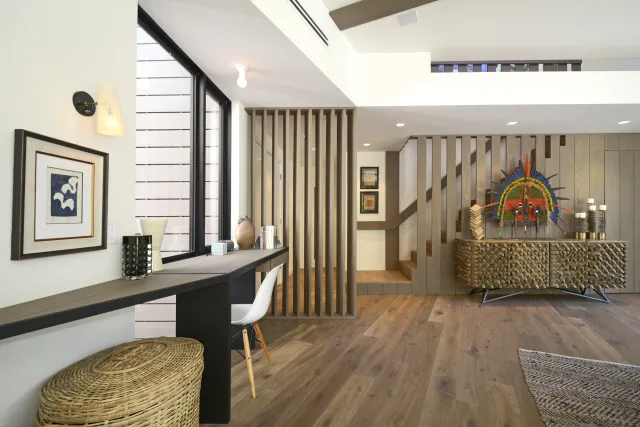 Modern living space with wood flooring, decorative cabinet, artwork, and a long desk with seating by a large window.