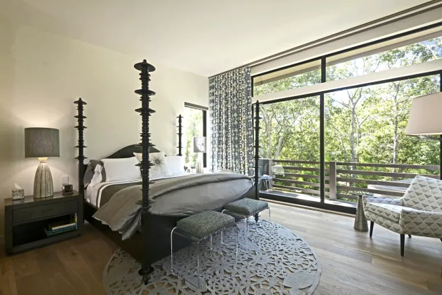 Modern bedroom with four-poster bed, patterned curtains, large windows, and scenic forest view outside.