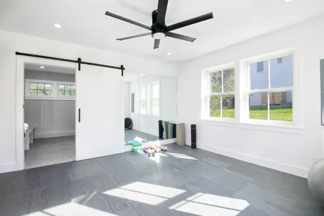 Bright home gym with large windows, sliding barn door, ceiling fan, yoga mats, and exercise equipment on gray flooring.